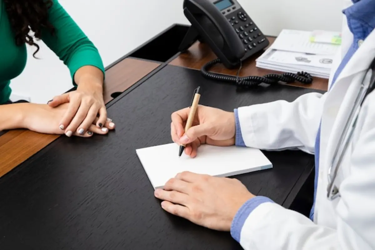 Médico anotando pedido em papel durante consulta, com paciente à mesa do consultório